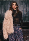 A woman models a fashion outfit featuring a light pink fluffy feather jacket draped over her shoulder, a sheer black long-sleeve top, and dark blue floral wide-leg pants, posed against a dark textured industrial background.