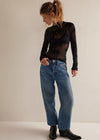 A woman wearing a sheer black long-sleeve top with a floral lace overlay, blue wide-leg jeans, and black ankle boots, standing against a plain off-white studio background.