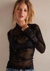 A woman wearing a sheer black long-sleeve top with floral lace applique over a black bralette, paired with blue jeans, standing against a plain beige wall.