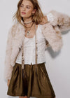 A woman models a light pink faux fur jacket over a white corset top and an olive green metallic skirt, styled with layered necklaces against a plain light gray background.