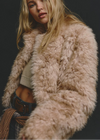 A close-up lifestyle shot of a model wearing a voluminous, light beige faux fur jacket over a beige crop top and blue jeans, set against a dark studio background.