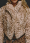 A close-up of a model wearing a cropped, shaggy faux fur jacket in a light beige and cream color, paired with a brown plaid skirt against a dark background.