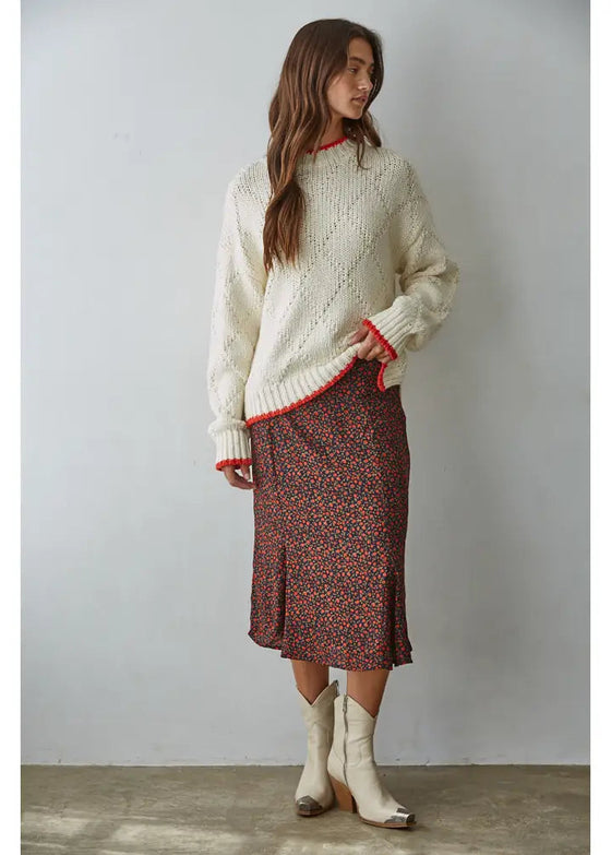 Woman wearing a cream sweater with red trim and a patterned skirt, standing against a plain background.