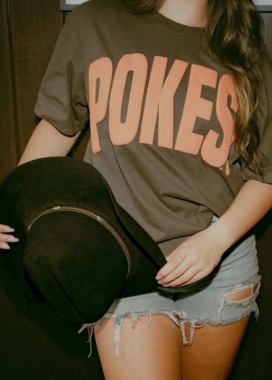 Person wearing a brown t-shirt with 'POKES' printed on it, holding a guitar case.