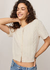 Woman wearing a beige cable knit cardigan and blue jeans against a plain background